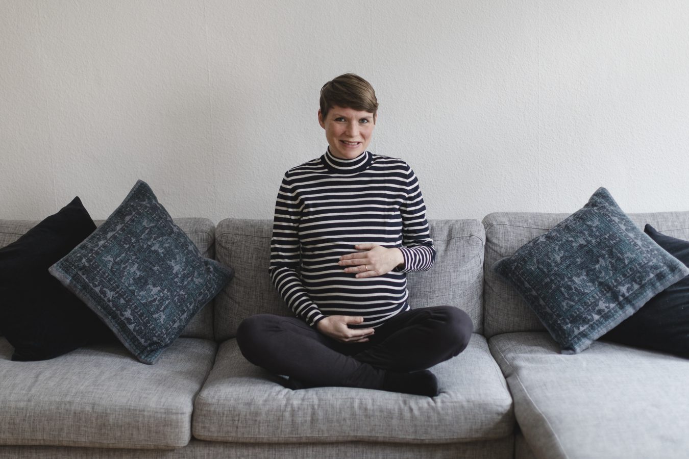 Babyfotograf Hamburg - Schwangere sitzt im Schneidersitz auf dem Sofa und lächelt in die Kamera