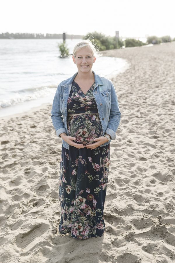 Schwangere steht im Sommerkleid am Strand